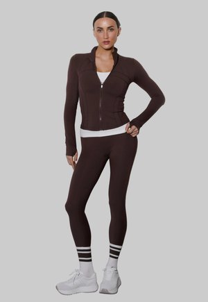 Kobieta ubrana w ciemnobrązową kurtkę na zamek i legginsy, biała koszulka na ramiączkach, paskowane skarpetki i białe trampki, stojąca na szarym tle.