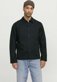 Zwarte denimjack met knoopsluiting, twee borstzakken en een kraag. Gemaakt met lichtblauwe jeans en een lichtbruine gebreide beanie.