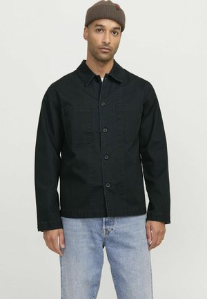 Black denim jacket with button closure, two chest pockets, and a collared neckline. Worn with light blue jeans and a brown knit beanie.