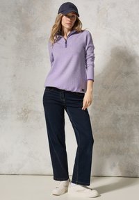 Helllila Zip-up-Pullover mit langen Ärmeln, dunkle Jeans im Straight-Leg-Schnitt und weiße Sneaker. Die Kappe hat ein schwarzes Logo-Detail.