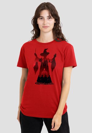 Woman wearing a red t-shirt featuring a black silhouette of a witch with a pointed hat and castle, holding a wand.