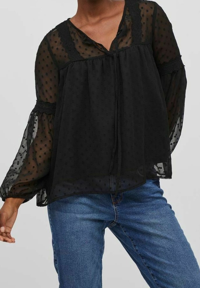 Černá průsvitná halenka s puntíkovaným vzorem, výstřihem do V a krajkovou volánkovou lemovkou. Má dlouhé, nafouklé rukávy a volný střih.