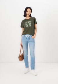 T-shirt vert olive avec un imprimé géométrique, jeans bleu clair, baskets blanches et sac à bandoulière marron. Look décontracté, tissu doux.
