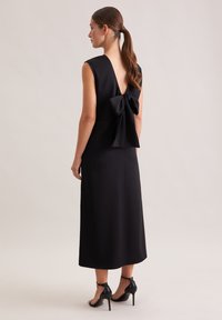 Robe noire sans manches avec un dos en V profond et un grand nœud à la taille. Tissu lisse avec une coupe élégante et ajustée. Talons aiguilles.