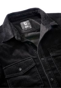 Chemise en velours côtelé noir avec un col classique, deux poches avant et des fermetures à boutons. Le tissu présente une texture côtelée. Étiquette de la marque visible.