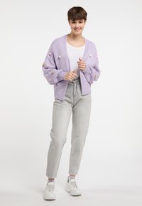 Lila ylisuuri neulecardigan, jossa on värikkäitä pompomeita, käytetty valkoisen paidan päällä, yhdistettynä vaaleanharmaisiin farkkuihin ja valkoisiin tennareihin.