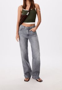 Jean large gris clair avec détails délavés, associé à un débardeur vert ajusté. Sac bandoulière marron texturé avec des accents métalliques. Sandales noires.
