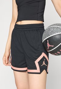 Shorts de sport noirs avec des accents roses, taille élastique, tissu texturé et détail de logo sur l'ourlet. Tenant un ballon de basket avec un design similaire.