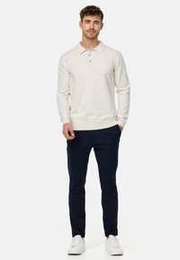 Svijetlosmeđi dugosleeve polo majica s tri gumba i rebrastim manžetama, uparen s tamnoplave uske hlače i bijelim tenisicama.
