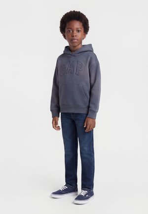 Αγόρι με σγουρά μαλλιά που φοράει ξεθωριασμένο μπλε φούτερ GAP, σκούρα μπλε τζιν και ναυτικά αθλητικά παπούτσια, στέκεται μπροστά σε ένα απλό ανοιχτό φόντο.