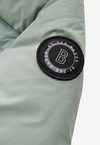 Lichtgroene stof met een ronde zwarte patch waarop een witte "B" staat en de tekst "By Ted Baker" rond de patch.