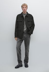 Donker wollen jas met voorkantenzakken, grijze trui eronder, gecombineerd met grijze spijkerbroek en zwarte leren schoenen, staand tegen een lichte achtergrond.