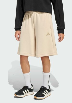 Shorts sportivi beige con vita elastica e tasche laterali, caratterizzati da sottili accenti con il logo Adidas. Indossati con scarpe da ginnastica nere e calzini bianchi.