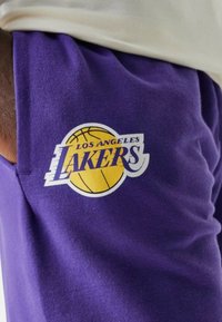 Pantaloni da tuta viola con il logo dei "Los Angeles Lakers". Il logo include un pallone da basket giallo ed è stampato sul tessuto.