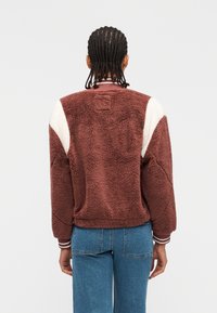 Άτομο με πλεξούδες που φοράει καφέ και λευκό χνουδωτό μπουφάν και μπλε τζιν, στέκεται γυρισμένο με την πλάτη προς απλό φόντο.