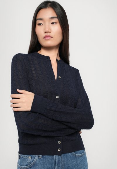 Tumši zila adīta kardigāna ar V formas kakla izgriezumu, kuru rotā teksturēts raksts, garām piedurknēm un trim pogām priekšā.
