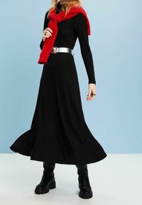 Langes schwarzes Kleid aus weichem Stoff mit langen Ärmeln und einem seitlichen Schlitz. Kombiniert mit einem leuchtend roten Schal und klobigen schwarzen Stiefeln. Silberner Gürtelakzent.