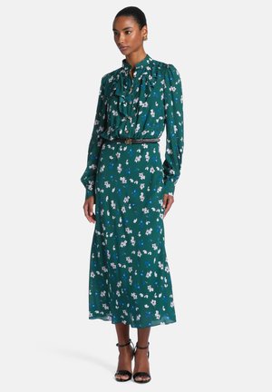 Femme portant une robe midi verte à manches longues fleurie avec un nœud noué au cou, une ceinture noire, des boucles d'oreilles dorées et des sandales noires à talons hauts.