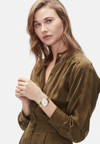 Goldene Armbanduhr mit einem weißen Zifferblatt, goldfarbener Band sowie metallischen Akzenten. Modell trägt eine olivgrüne Bluse mit Knopfleiste und weicher Textur.