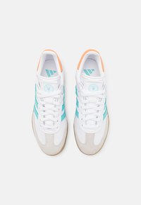 Zapatillas deportivas blancas con rayas turquesa y acentos naranjas, que presentan un upper sintético liso y una puntera de ante beige texturizado.