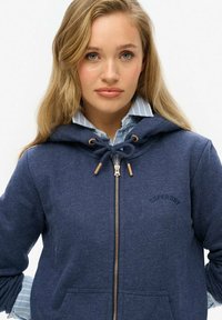 Sudadera con cremallera color navy hecha de tela suave, con capucha con cordón, bolsillos frontales y un discreto bordado de logo. Camiseta a rayas debajo.