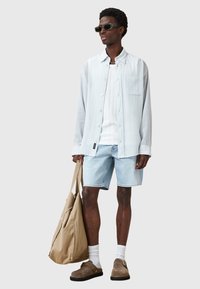 Camicia a righe blu chiaro con bottoni sopra una canotta bianca, abbinata a pantaloncini di denim chiaro, scarpe slip-on marroni e una borsa tote beige.