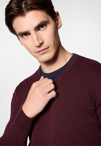 Burgundowy sweter z dekoltem w serek o teksturowanym wzorze, noszony na ciemnoniebieskiej koszuli. Model dotyka kołnierza, podkreślając detale i dopasowanie.