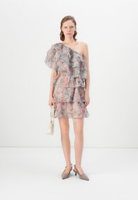 Kvinna som står i enaxlad lagerblommig klänning, håller en liten vit handväska, bär spetsskor i taupe färg, enkel vit bakgrund.