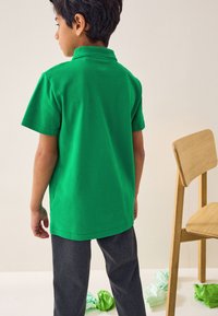 Žalia trumpomis rankovėmis polo marškinėliai su apykakle, derinami su tamsiai pilkomis kelnėmis. Fone matoma šviesi, neutrali spalva.