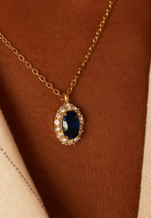 Collier en chaîne dorée avec un pendentif en pierre bleue ovale, entouré de petites pierres claires, sur une peau brune texturée, partiellement recouverte d'un tissu crème.