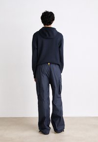 Personne aux cheveux bouclés et foncés se tenant face à un mur blanc, portant un sweat à capuche bleu marine et un pantalon cargo bleu marine ample avec poches latérales et chevilles élastiques.