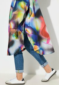 Abrigo largo colorido y brillante con patrones abstractos, combinado con jeans de mezclilla remangados y zapatillas blancas con detalles negros.