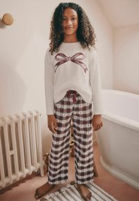 Vit långärmad skjorta med en röd rosettgrafik, ihop med rutiga byxor i rött, grönt och vitt med små hjärtan. Mjukt tyg, avslappnad passform.