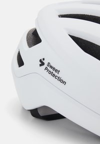 Casque de vélo blanc avec des accents noirs et des ouvertures de ventilation, arborant le logo "Sweet Protection" sur le côté. Design lisse et léger.