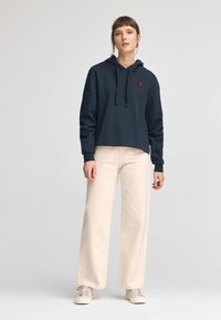 Marinblå hoodie med dragsko och en logotyp på bröstet, kombinerat med ljusbeiga vida byxor och beiga sneakers. Slät tygstruktur.