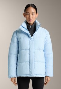 Femme portant une doudoune bleu clair à fines rayures sur un haut noir zippé, debout devant un fond beige uni.