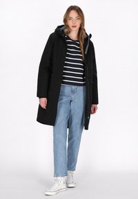 Lungo cappotto nero con cappuccio, abbinato a una maglietta a righe blu navy e bianco, jeans azzurri chiari e sneakers bianche con accenti colorati.