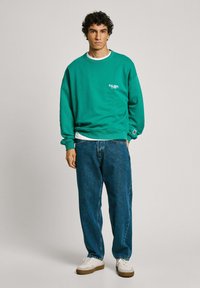 Groen sweatshirt met ribbelmanchetten en ronde hals, met een klein logo. Gecombineerd met losse blauwe denim jeans en witte sneakers.
