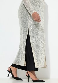 Largo vestido plateado con lentejuelas y una profunda abertura lateral, combinado con elegantes tacones negros. Textura suave y diseño ceñido.