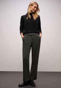 Czarny sweter z wysokim kołnierzem, zestawiony z szarymi spodniami o teksturze i czarnymi botkami za kostkę. Model stoi swobodnie z rękami w kieszeniach.