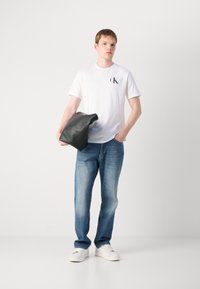 Bijela pamučna majica s crnim logom, plave traperice i bijeli tenisice. Model drži crnu kožnu torbu. Jednostavan i ležeran dizajn.