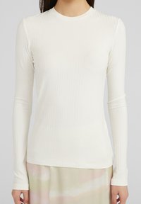 Kvinna klädd i en åtsittande, långärmad ribbad vit topp och en ljus kjol med mjuka pastellmönster.