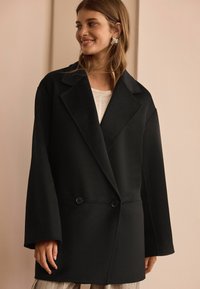 Svart ullblandad kappa med överdimensionerade slag, dubbelknapprad och vida ärmar, som har en elegant, strukturerad silhuett.