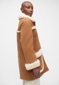 Abrigo exterior marrón con un suave cuello y puños de borrego crema, que presenta un corte recto y cierre de botones. Textura suave y líneas limpias.