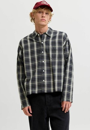 Jeune homme aux cheveux blonds bouclés portant une chemise à carreaux foncée boutonnée, un pantalon noir et une casquette de baseball rouge, debout devant un fond uni.