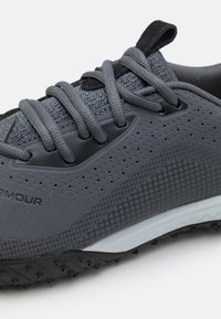 Zapatilla deportiva gris con parte superior sintética texturizada, diseño perforado y cordones a juego. Cuenta con una suela de goma duradera con pequeñas protuberancias de tracción.