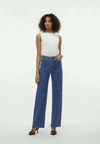 Vysokoklasové modré džínsy so širokými nohavicami, zdobené striebornými cvočkami. Skombinované s priliehajúcim bielym topom bez rukávov a pruhovanými podpätkami.