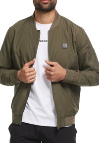 Homme portant une veste bomber vert olive en train de la déboutonner, par-dessus un t-shirt blanc avec un texte noir, sur un fond blanc uni.