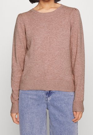 Personne portant un pull en tricot à manches longues de couleur marron clair associé à un jean en denim bleu clair, debout devant un fond uni.