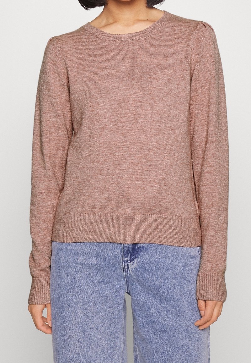 Personne portant un pull en tricot à manches longues de couleur marron clair associé à un jean en denim bleu clair, debout devant un fond uni.